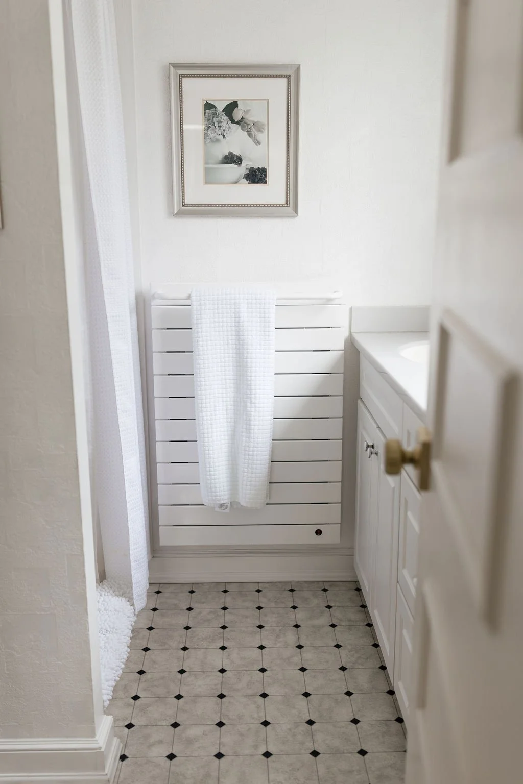 A small, white bathroom with a closed door, a white sink cabinet, a towel rack with a white towel, a framed black and white floral artwork, a black and white checkered tile floor, and a white radiator.