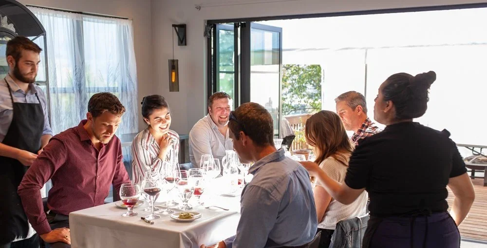 A group of people sitting around a dinner table, enjoying a meal and drinks in a bright, modern restaurant or home dining room.