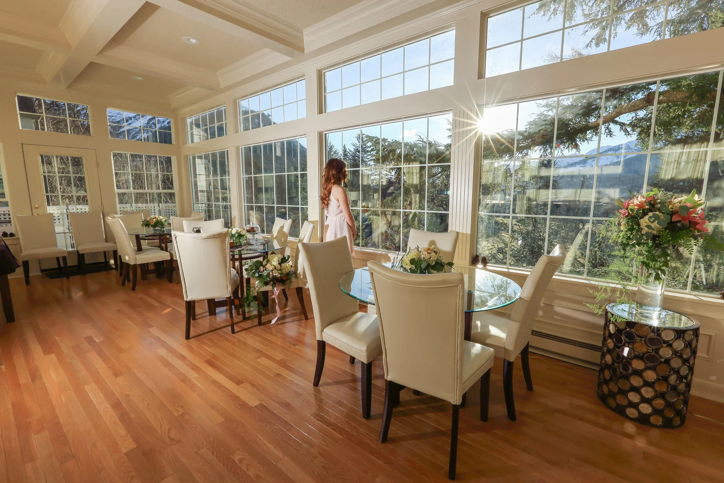 Sunlight streaming through large glass windows into a dining room with white chairs, a glass table, and flower arrangements, with a woman standing near the windows.
