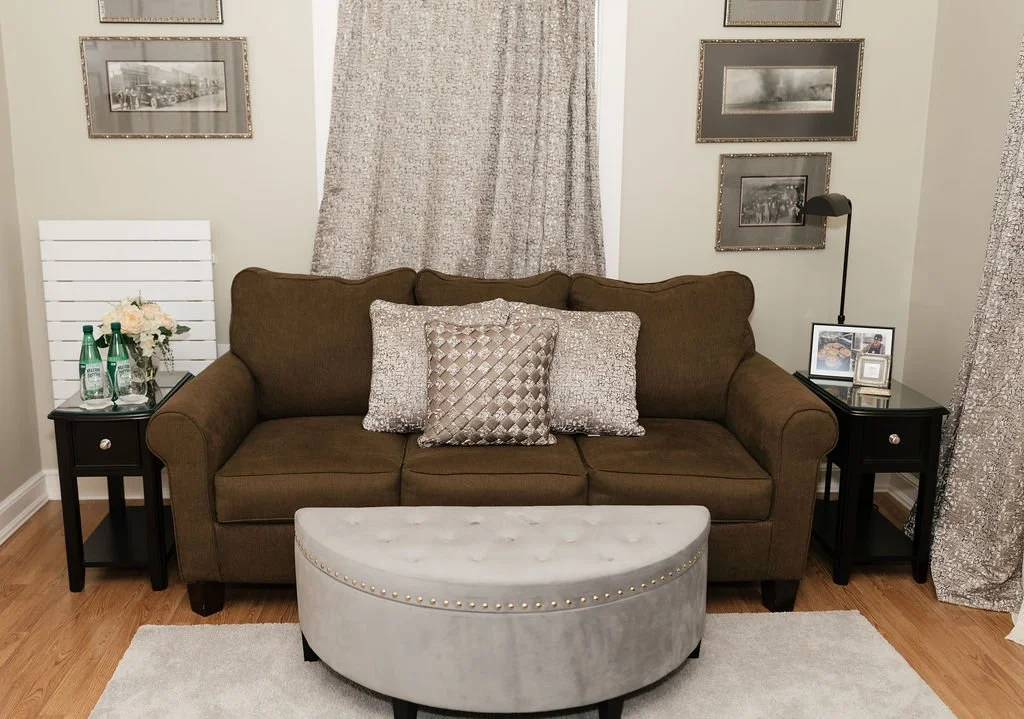 Living room featuring a brown couch with three decorative pillows, two black side tables with decor items, wall art, beige curtains, and a white ottoman in front.