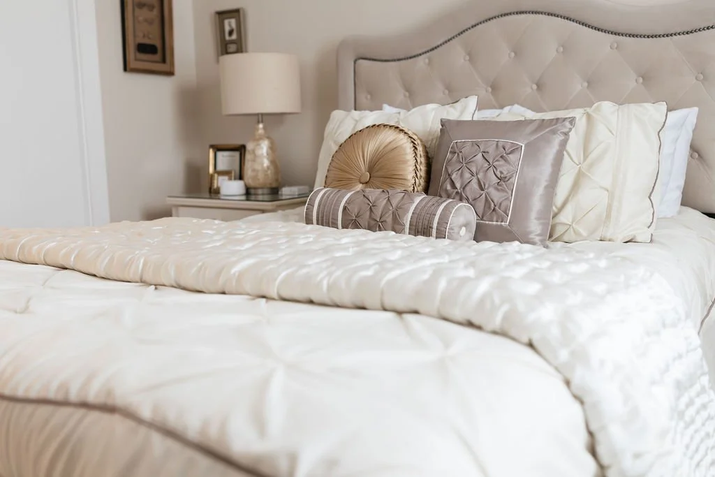 A neatly made bed with a beige tufted headboard, cream and beige pillows, including a round decorative pillow, in a room with a nightstand, lamp, and framed pictures.