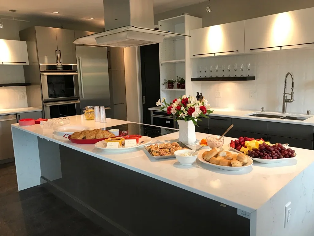 Modern kitchen with a white island countertop holding a variety of foods including fresh fruit, bread rolls, muffins, a large cake, and flowers in a vase. There are cups, plates, and utensils on the counter as well.