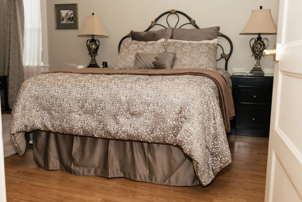 A bedroom with a bed featuring a metallic and beige bedspread, multiple pillows, and dark nightstands on each side with lamps. Wallpaper and framed picture seen in the background.