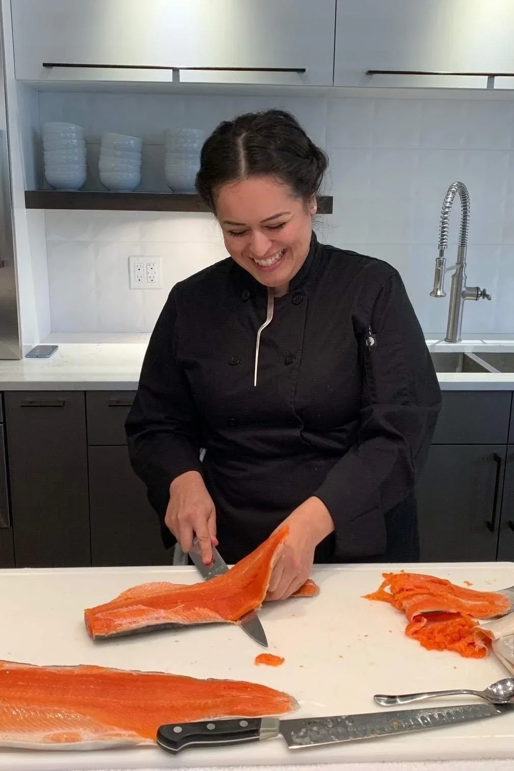 A woman in a black chef's coat preparing a large fillet of salmon in a modern kitchen, smiling as she cuts the fish on a white countertop.