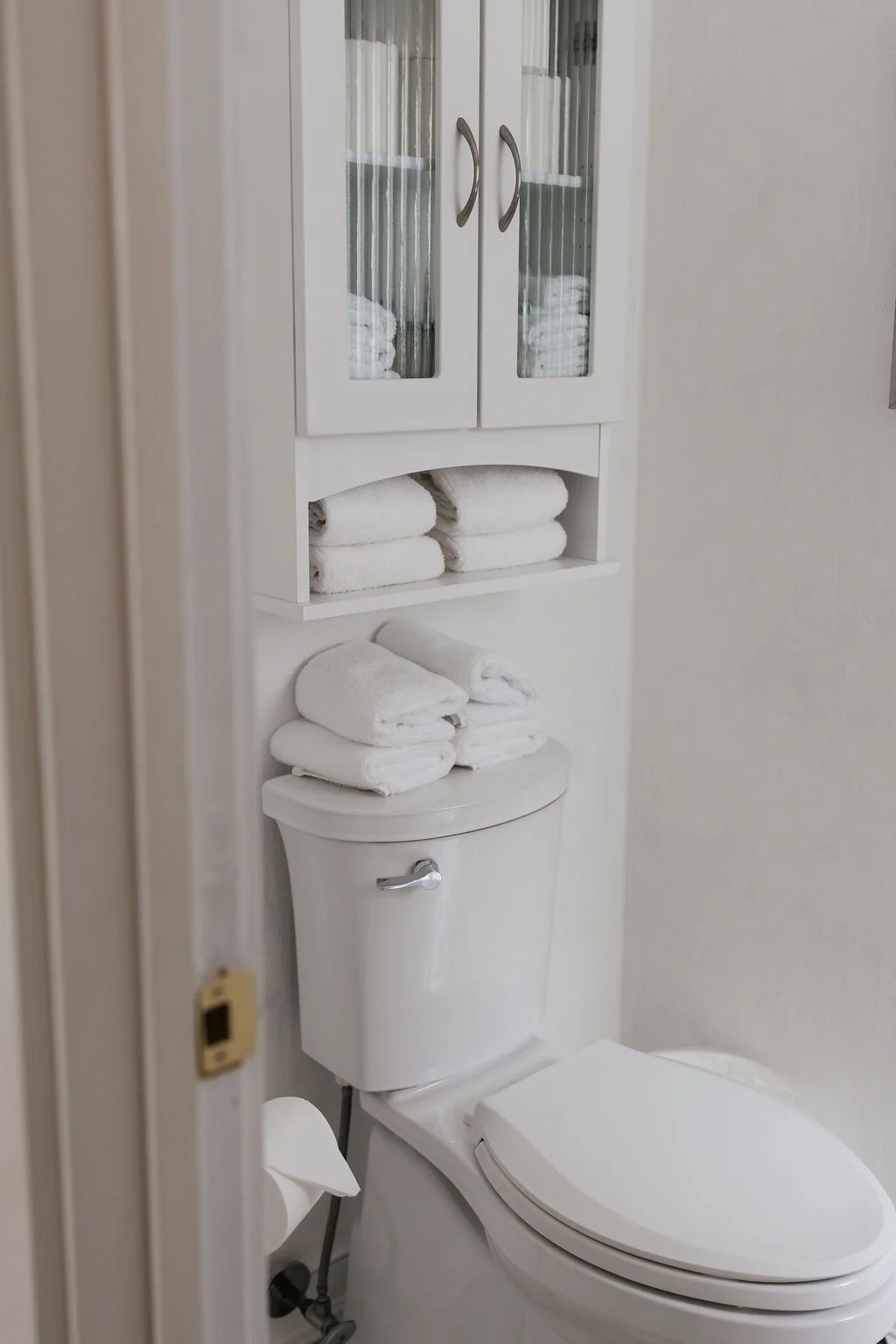Bathroom with a white toilet, and a white cabinet with two glass doors containing towels, and additional folded towels stored above the toilet.