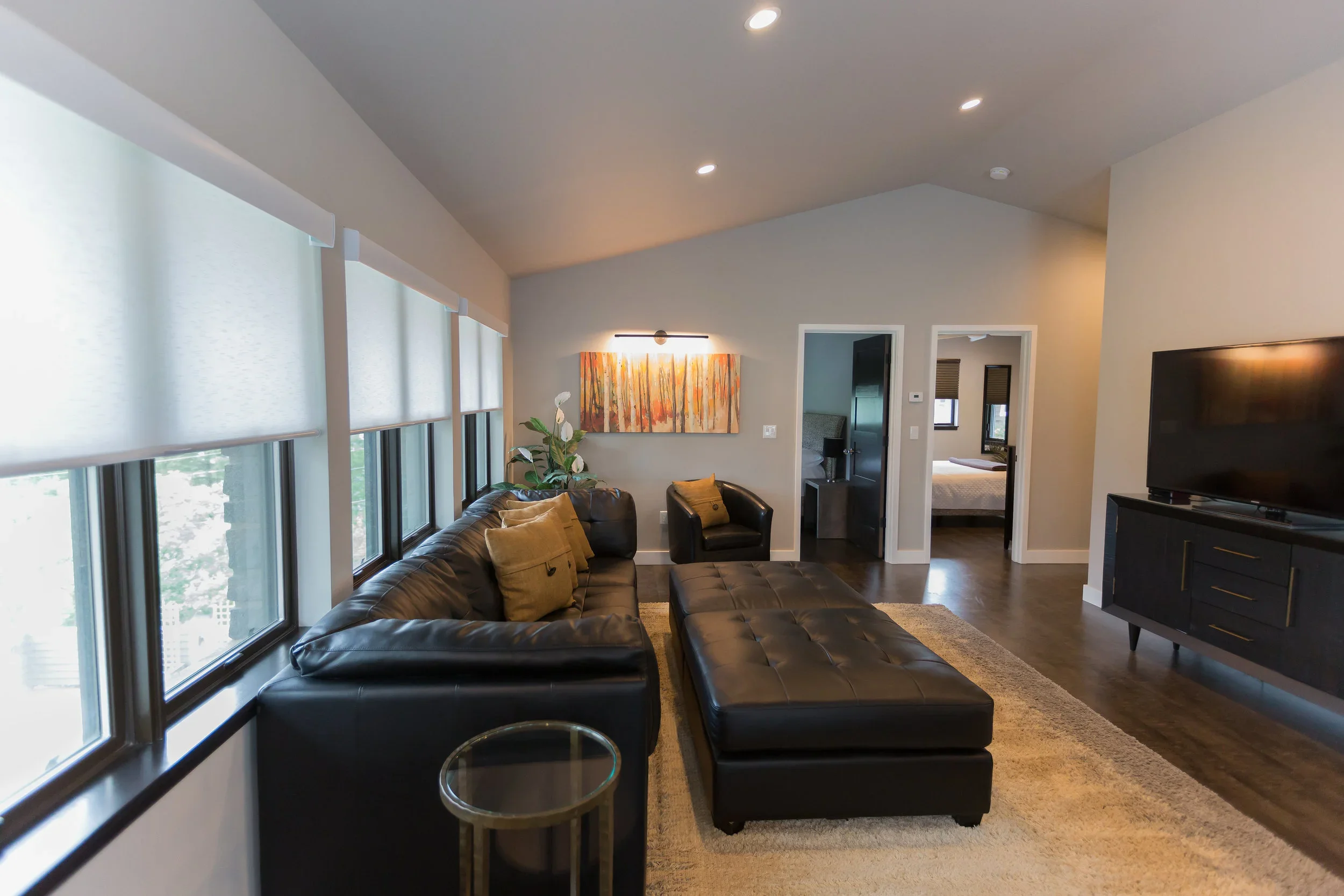 Living room with large window with white blinds, black leather sectional sofa with beige pillows, a small round glass side table, a tufted leather ottoman, and a wall painting with warm colors, adjacent to a doorway leading to another room with a bed