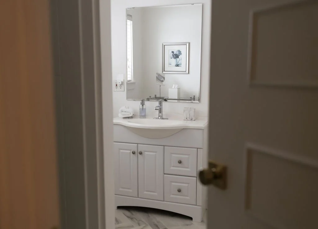 A bathroom sink with a mirror, a framed picture, and a window, viewed through an open door.
