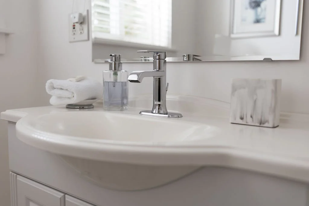 A bathroom sink with a chrome faucet, a soap dispenser, a folded towel, and a decorative box on the countertop, with a mirror and window in the background.