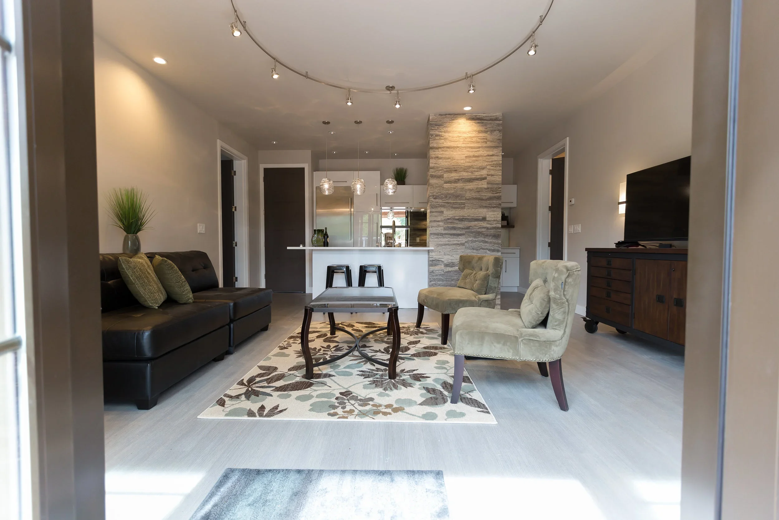 Living room with a black sofa, beige armchairs around a coffee table, a patterned rug, and a large flat-screen TV on a wooden stand. The room has a kitchen in the background with pendant lighting, white cabinets, and a stone accent wall.