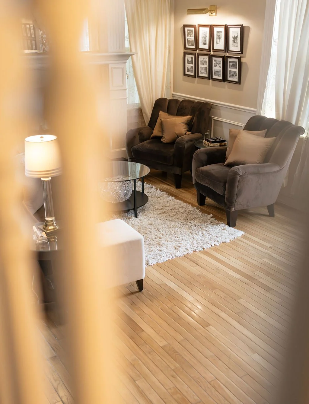 Living room with two gray armchairs, a small round coffee table, a table lamp, a framed photo collage on the wall, and cream-colored curtains.