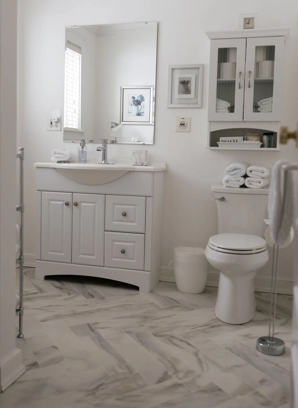 A clean bathroom with a white vanity, mirror, and cabinet; folded towels on the toilet tank; framed artwork and a window with blinds; chrome fixtures; marble flooring.
