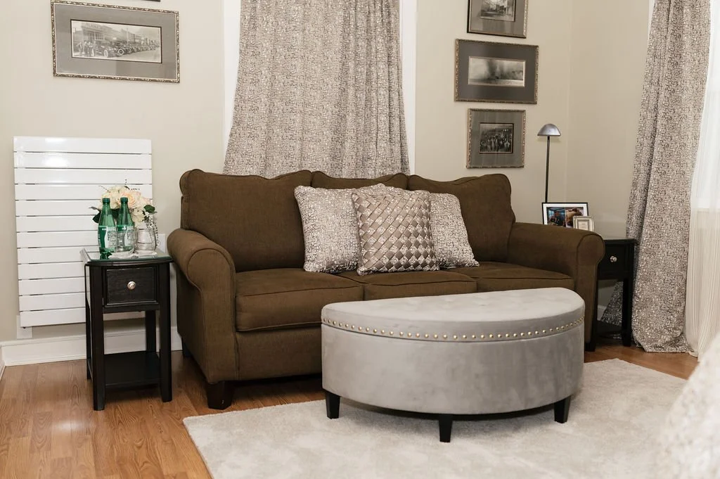 Living room with a brown couch, beige and patterned pillows, a gray ottoman, two black side tables with lamps, framed photographs on the wall, curtains, and a white radiator.
