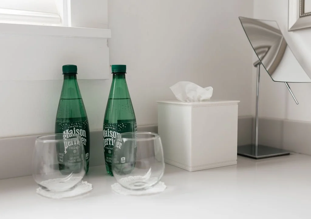 Two green bottles of Perrier sparkling water, two empty glasses on white coasters, a white tissue box, and a modern silver lamp on a white surface near a window.