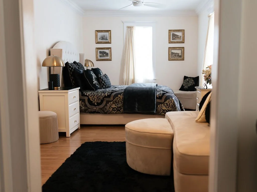 A bedroom with a bed, nightstands, framed pictures on the wall, and a bench at the foot of the bed. There is a black and gold color scheme with cream furniture and hardwood floors.