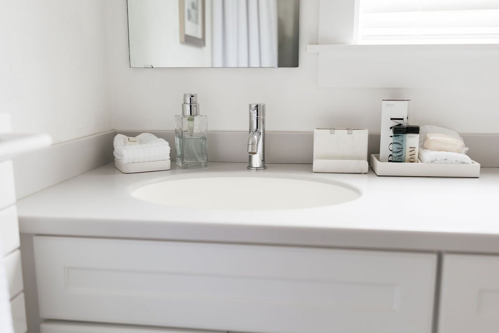 A white bathroom sink with a faucet, soap dispenser, folded towels, and toiletries on the countertop, with a mirror and window in the background.