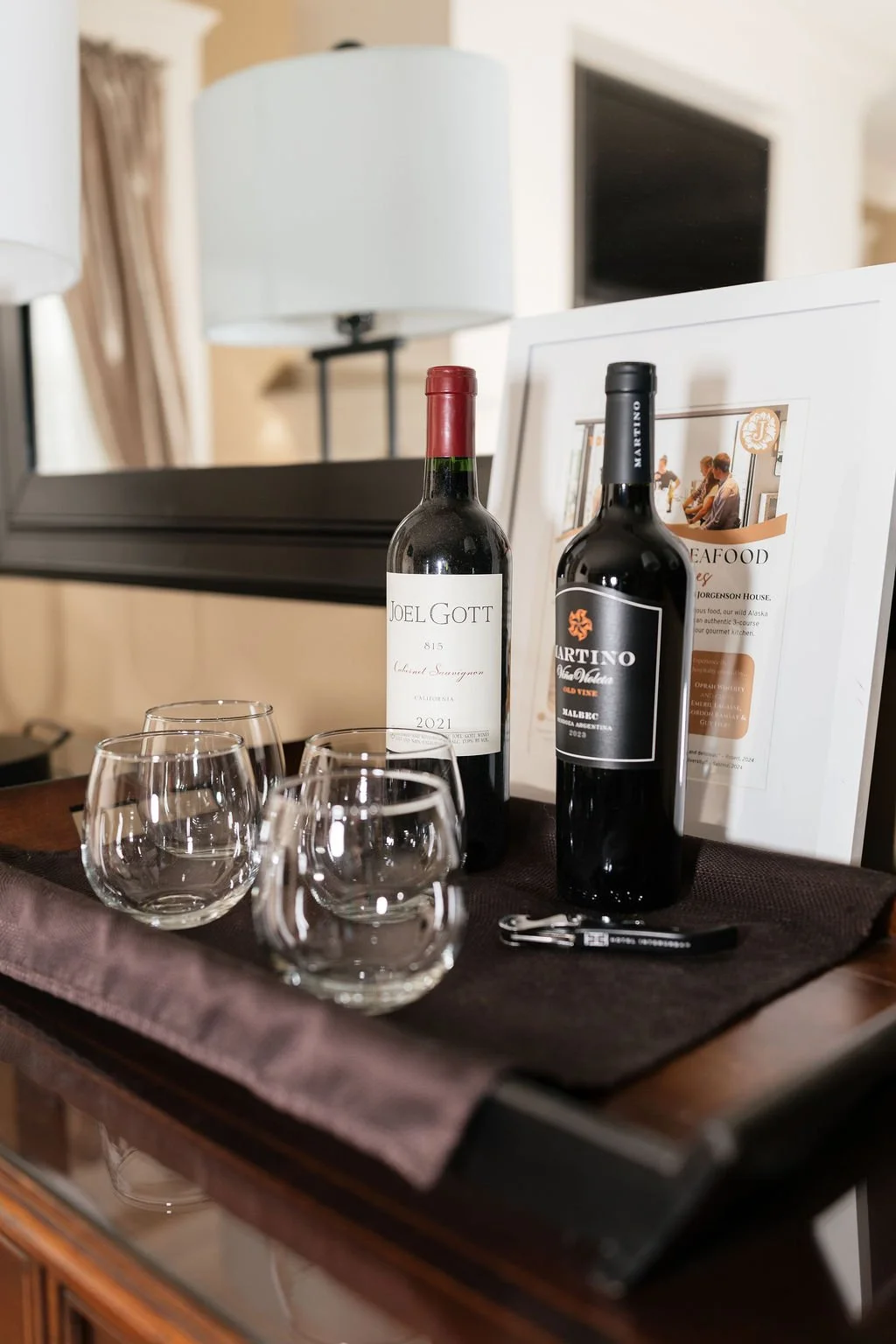Two bottles of red wine, three empty wine glasses, a black wine opener, and a framed sheet of paper, all arranged on a dark table runner on a wooden surface with a mirror and a lamp in the background.