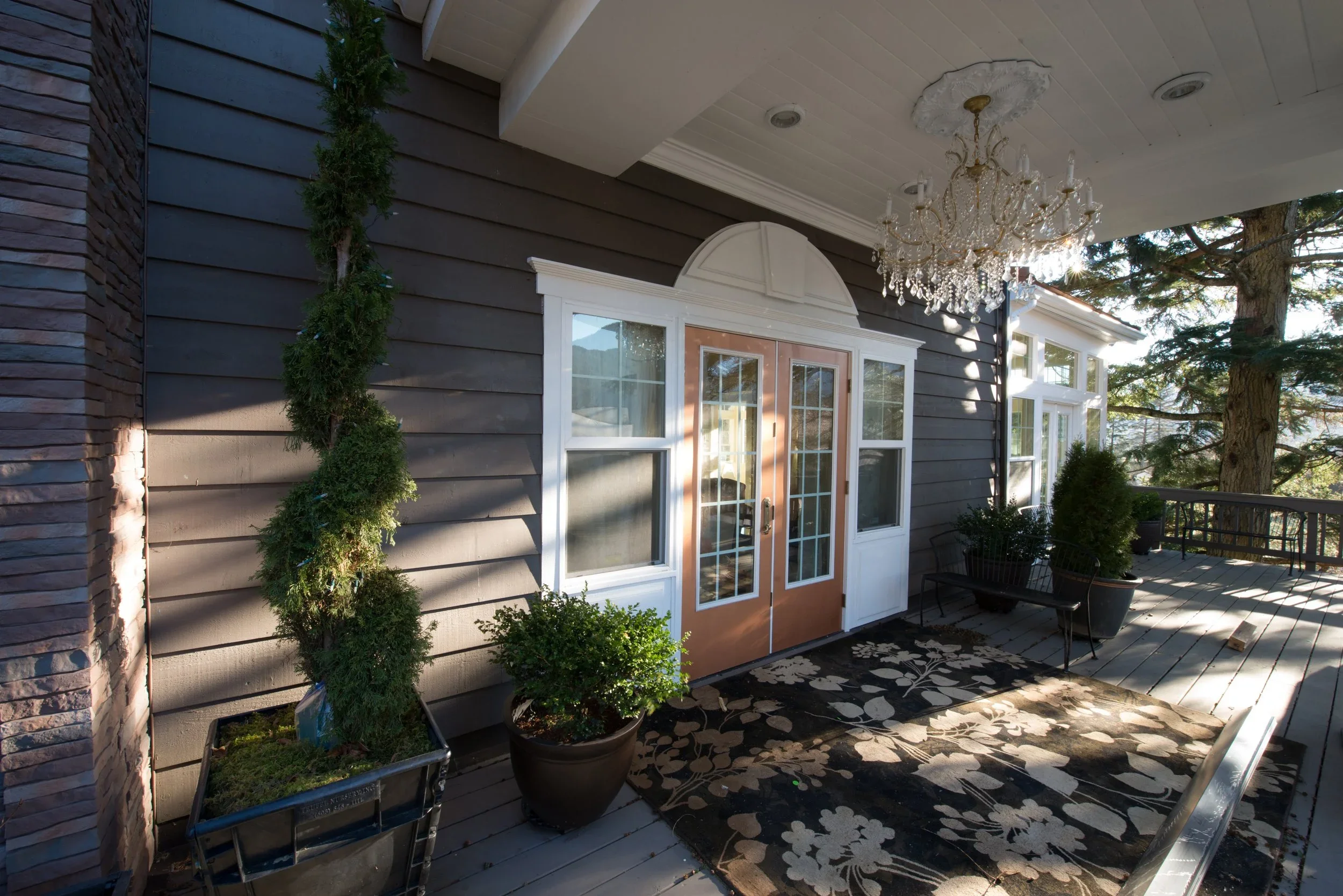 A covered porch area of a house features a chandelier hanging from the ceiling, potted plants, a rug, and a door with glass panels. There are trees and other houses visible in the background.