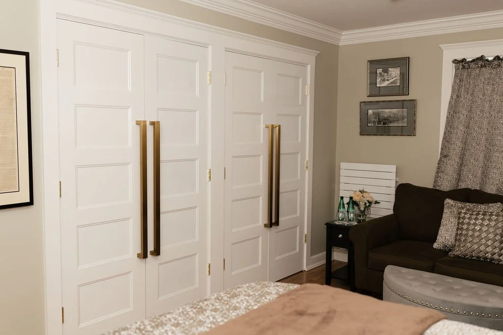 A closet with white double doors and gold handles, adjacent to a clinical style black side table with glasses and bottles, and a dark gray sofa with patterned cushions in a neutrally decorated room.