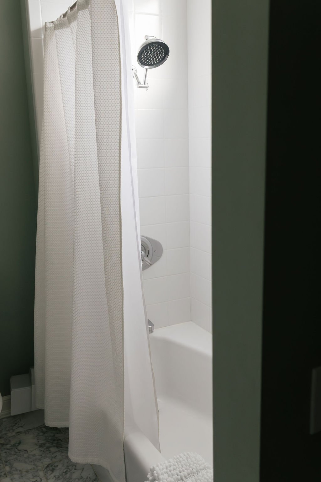 View of a shower stall with a white curtain, a handheld showerhead, and white tiled walls.
