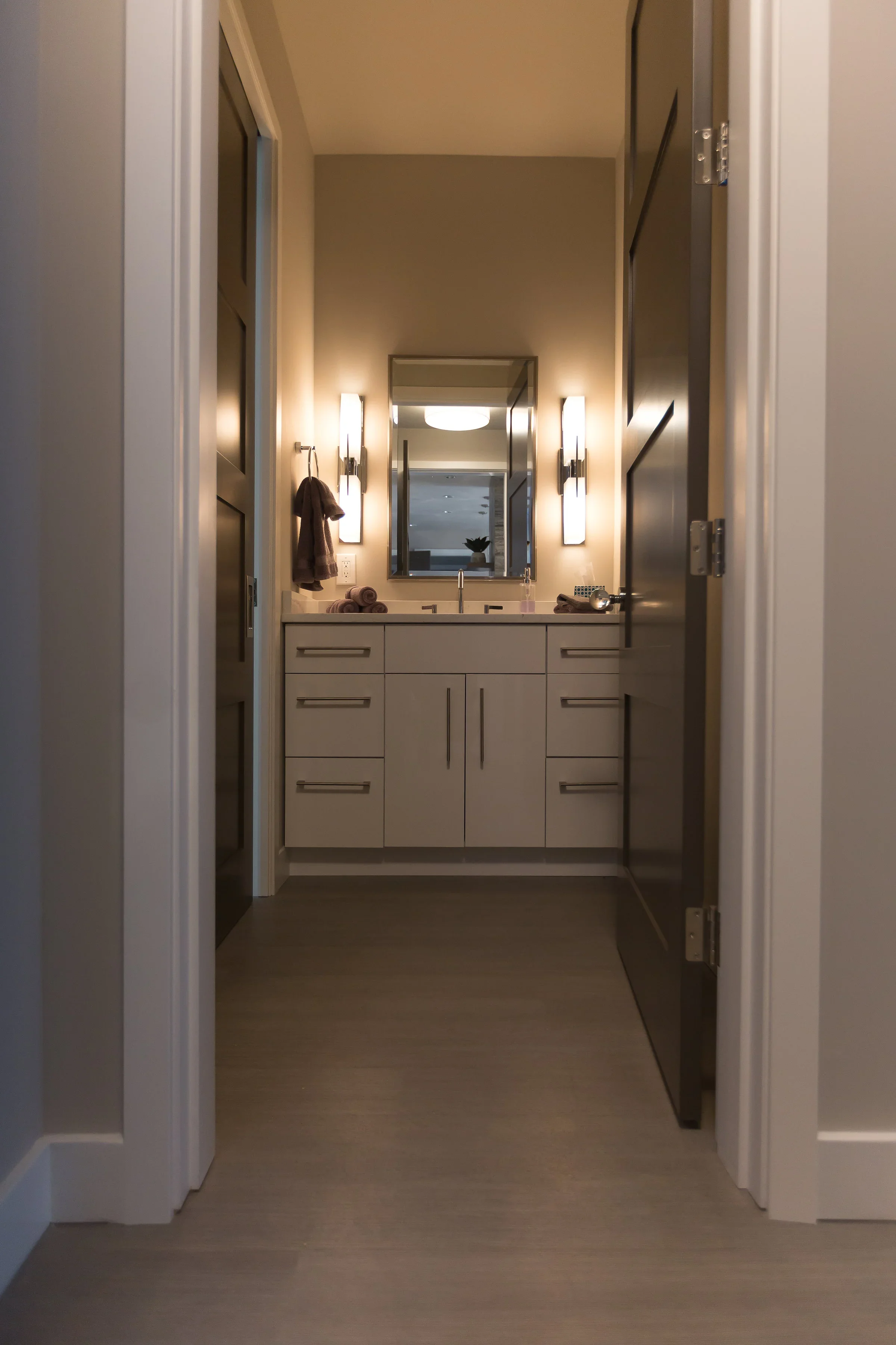 A bathroom vanity with a mirror, two wall-mounted lights, a brown towel hanging, and rolled towels on the counter, viewed through an open door in a modern room.
