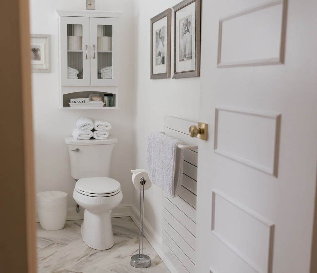 A small, white bathroom with a closed door, framed artwork on the wall, a toilet with rolled towels on the back tank, a white cabinet with glass doors, a trash can, a towel holder with a towel, and a toilet paper stand.