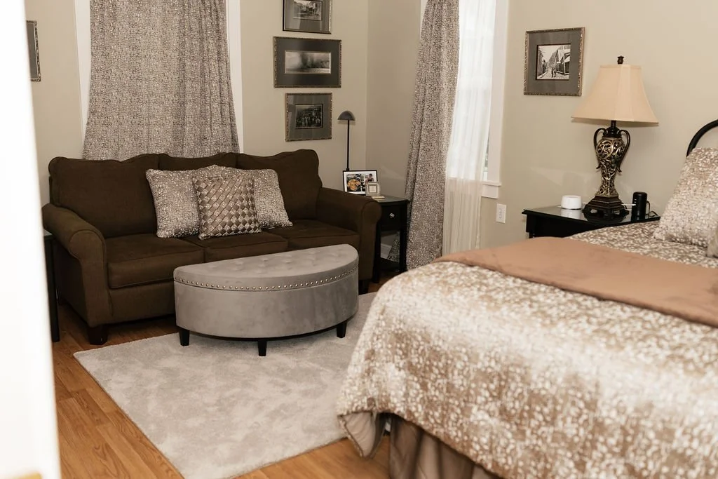 A cozy bedroom corner with a brown couch, decorative pillows, a gray ottoman, a black nightstand with photos and a lamp, a bed with beige patterned bedding, a nightstand with a lamp, and framed photos on the walls.