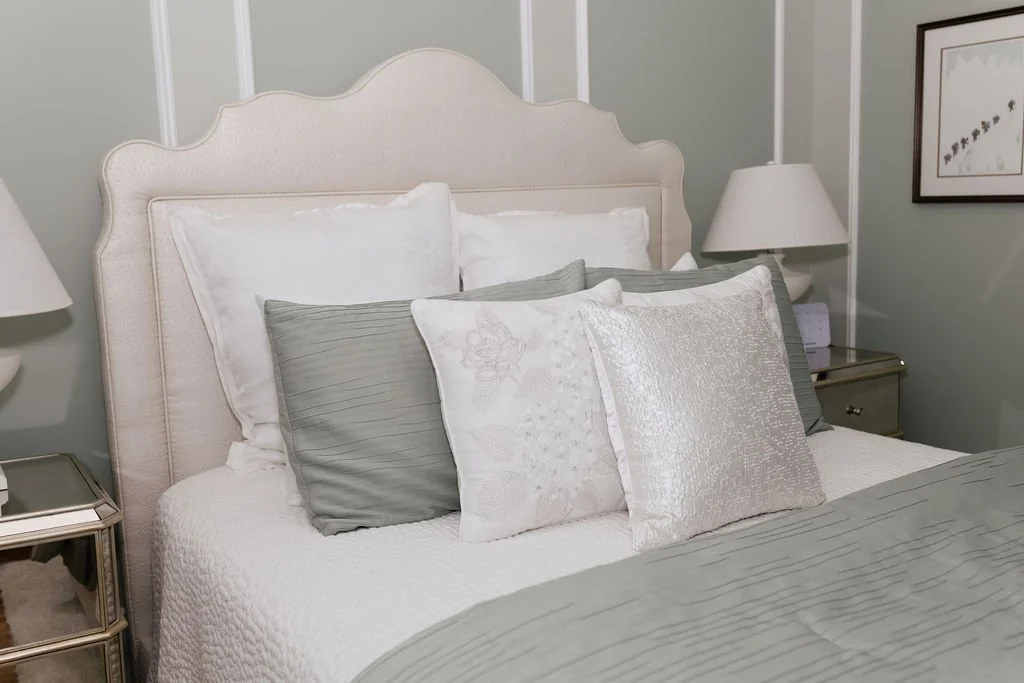 A neatly made bed with multiple pillows in a bedroom, flanked by two bedside tables with lamps, and a framed picture hanging on the wall.