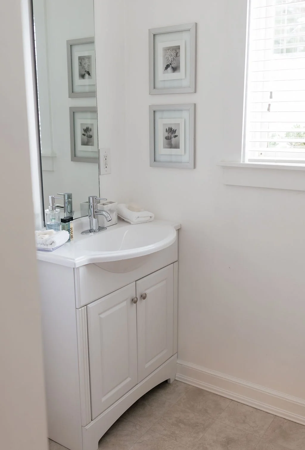 A small white bathroom vanity with a sink, a chrome faucet, a mirror, and framed black-and-white botanical photographs on the wall. There are toiletries and towels on the vanity, and a window with white blinds.