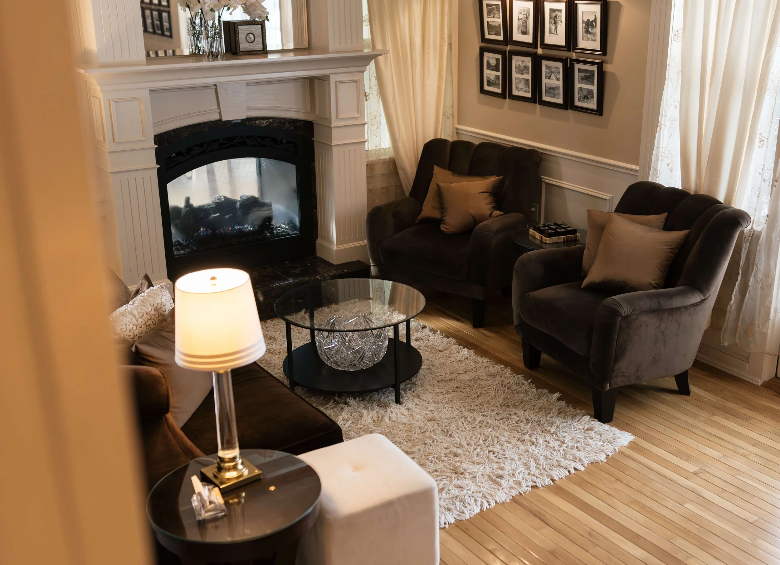 A cozy living room features a white fireplace with a glass cover, a black armchair with beige pillows, a plush white rug, a round black coffee table with a decorative glass item on top, and framed black and white photographs hanging on the beige walls, with large windows with sheer curtains allowing natural light.