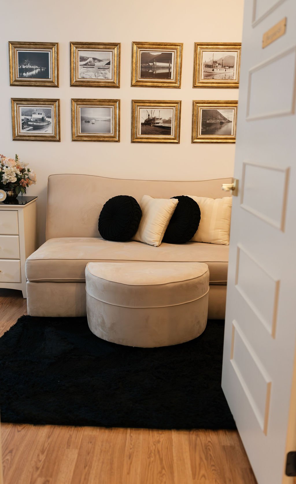 View of a cream-colored sofa with two black pillows and two cream pillows, a black rug underneath, a round ottoman in front, a white side table with a flower arrangement, and framed black and white photographs of ships on the wall in a cozy room.