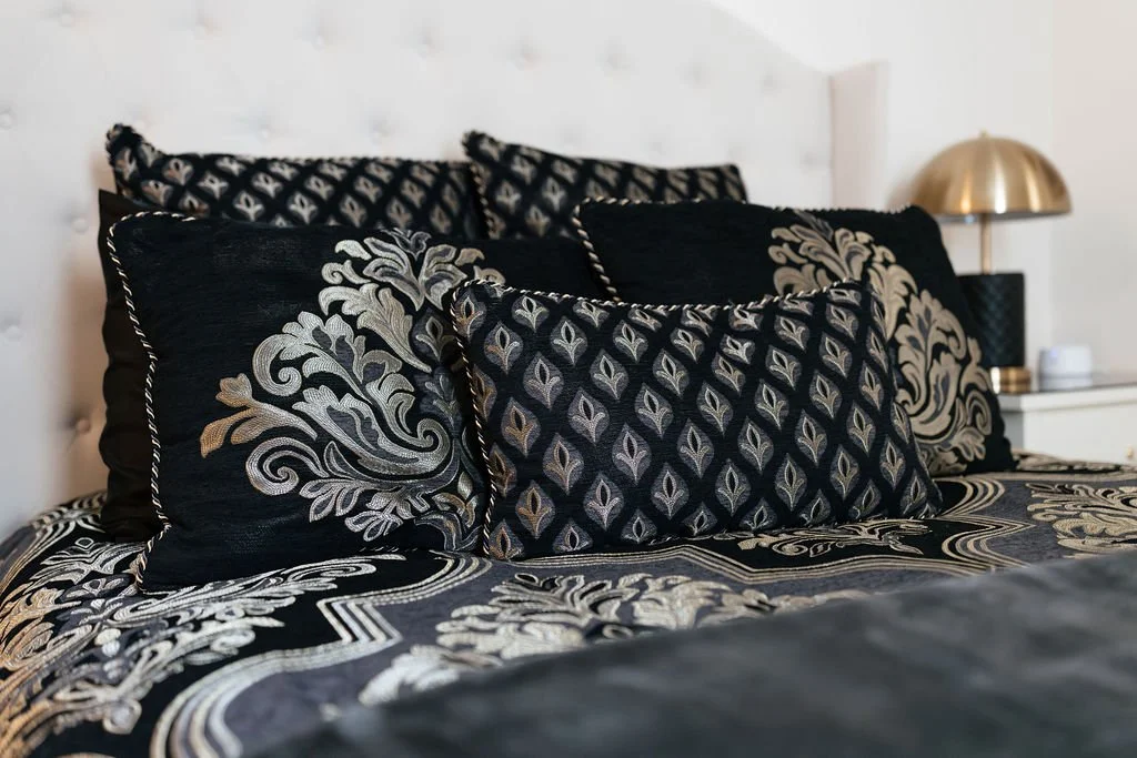A bed with black and gold patterned pillows and matching bedding, a white headboard, and a nightstand with a gold and black lamp in a bedroom.