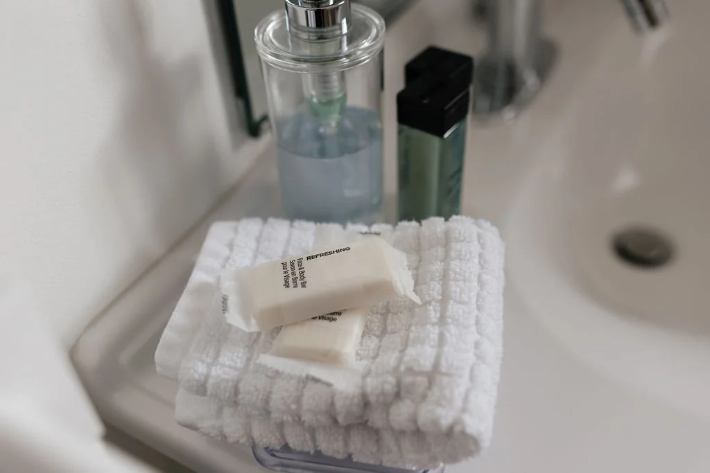 A white towel with a small white soap bar on top, placed on a surface next to a clear liquid soap dispenser, a black and green container, and a white sink in the background.