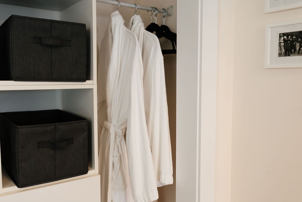 Two white bathrobes hanging in a closet with black storage bins on a white shelf to the left.