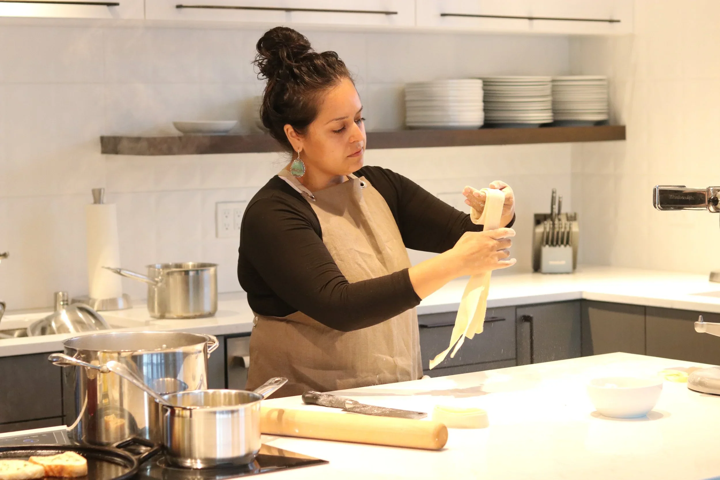 A woman in a kitchen preparing pasta, holding a long strip of dough.