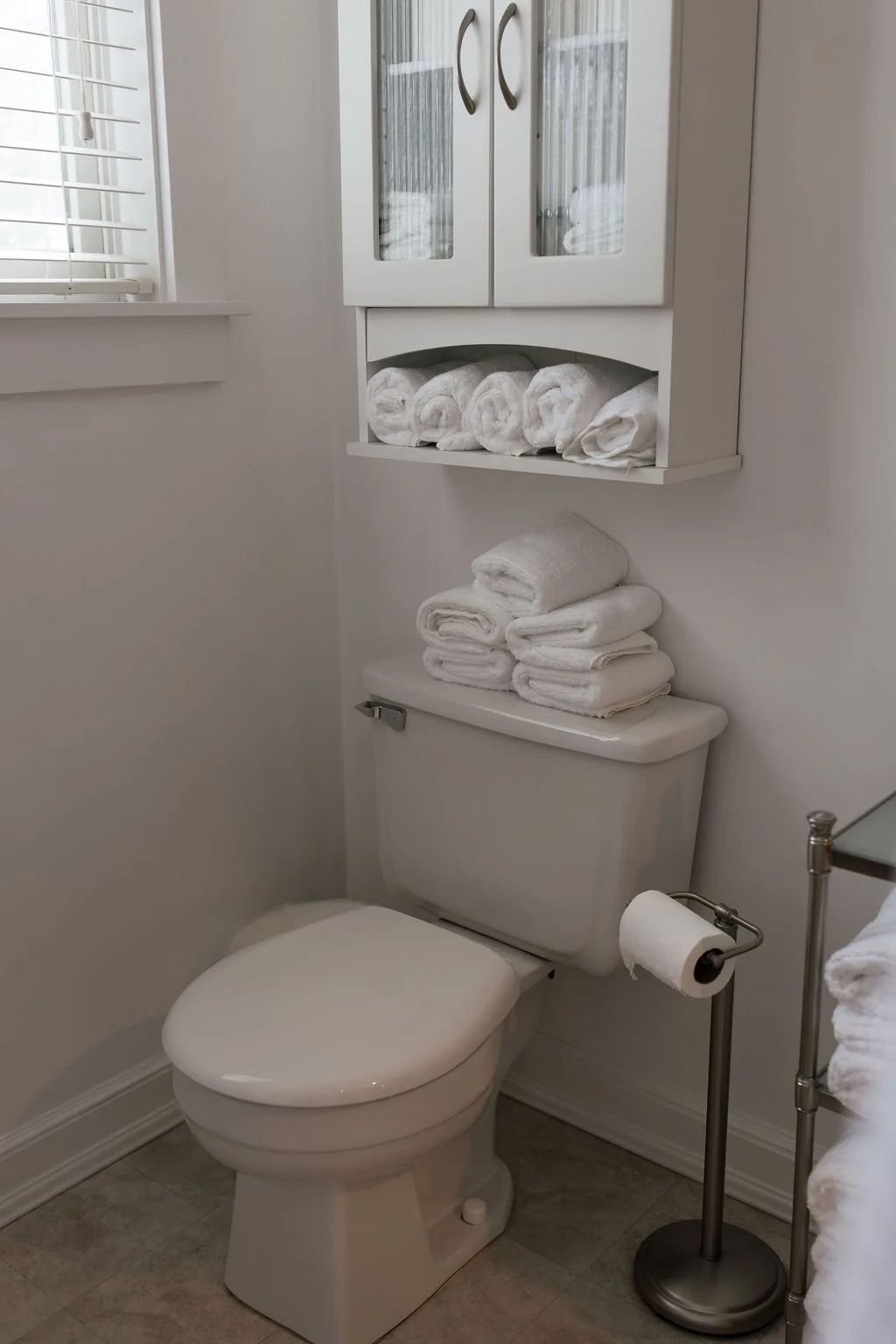 A white bathroom with a toilet, a wall-mounted cabinet with glass doors filled with rolled white towels, a shelf with folded white towels, and a metal toilet paper holder with a roll of toilet paper.