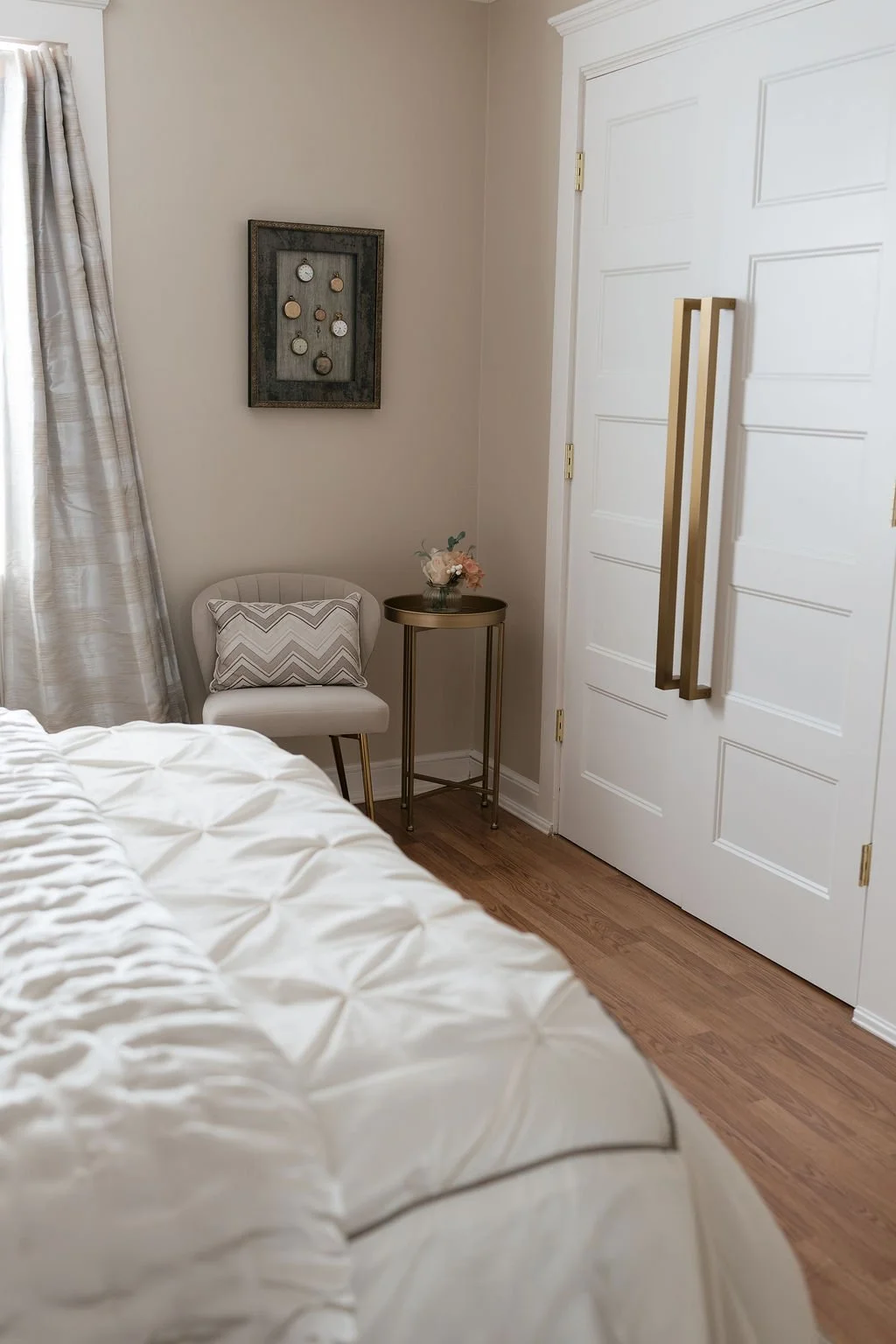 Corner of a bedroom with a bed, a white chair with a patterned pillow, a gold side table with a flower arrangement, a framed hanging jewelry display with watches, and a white closet door with gold handles.