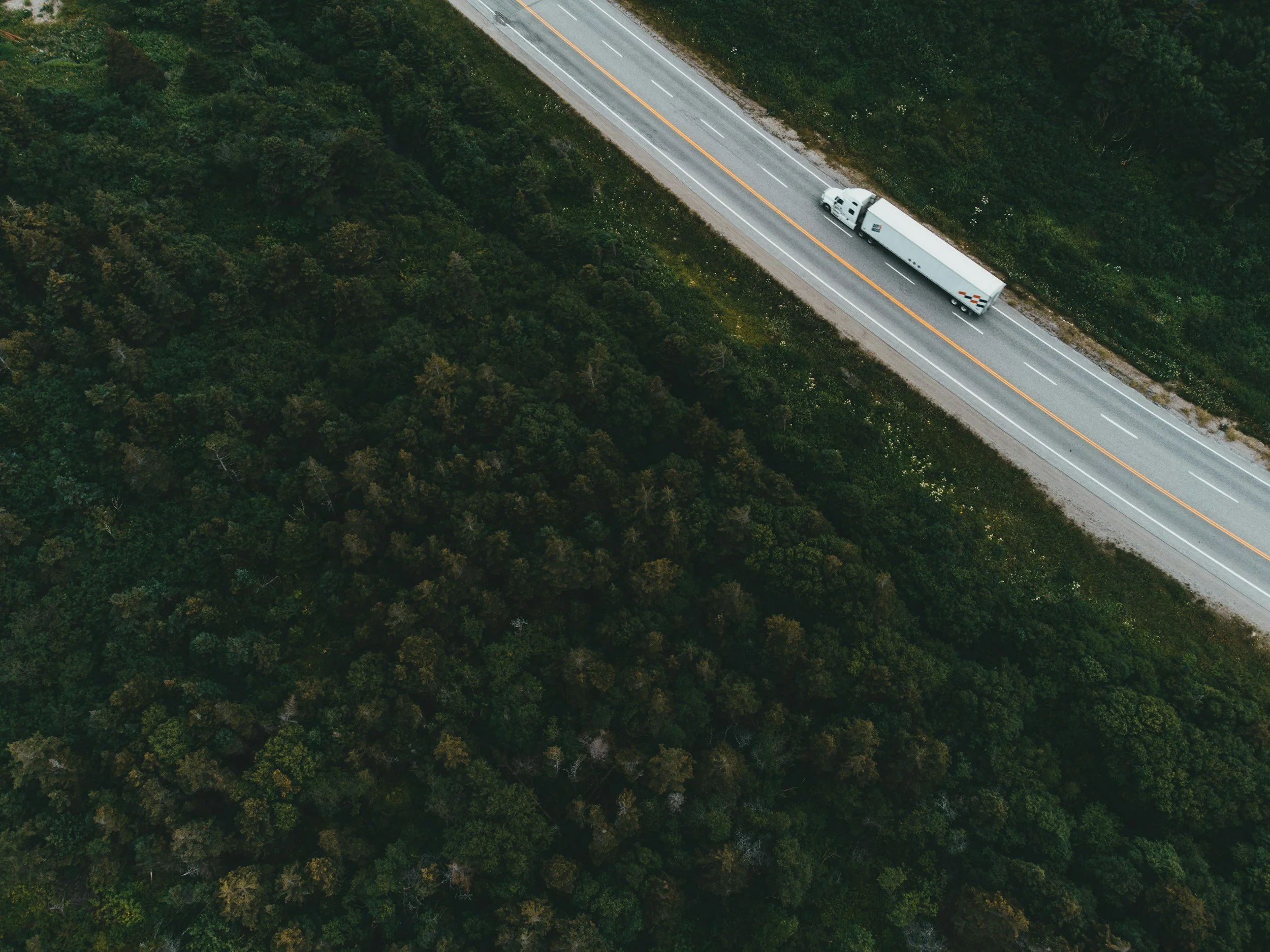 An aerial view of a highway running through a forest with a large semi-trailer truck traveling along it.