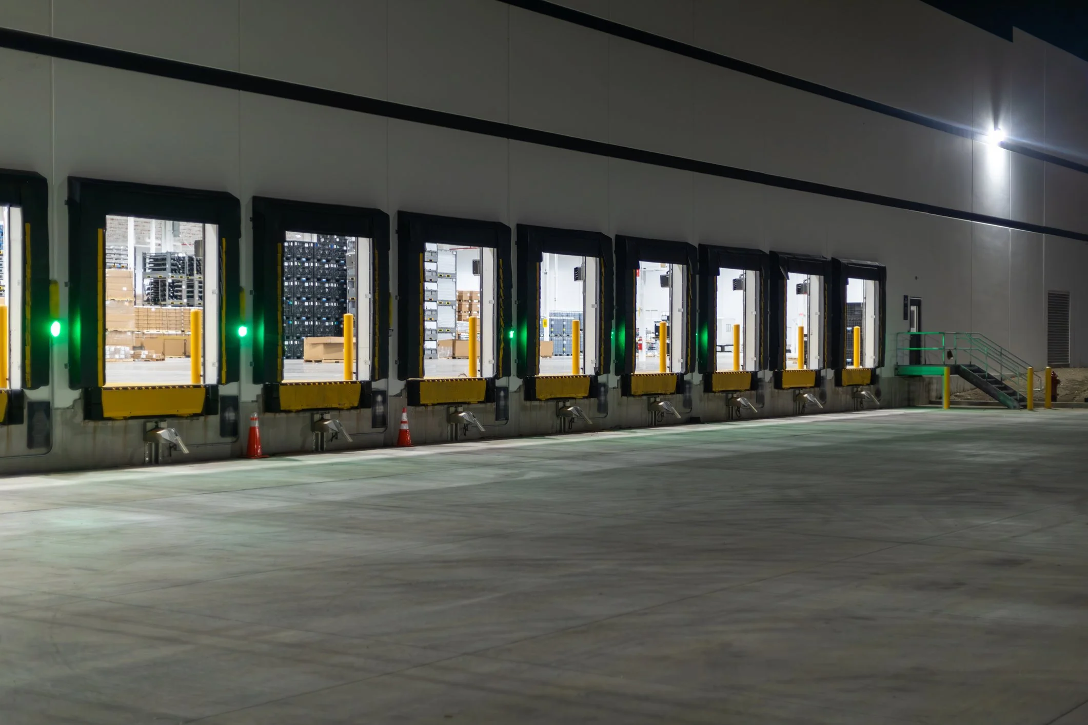 Loading dock with multiple automated truck doors, illuminated with green lights, and surrounded by safety cones and barriers.