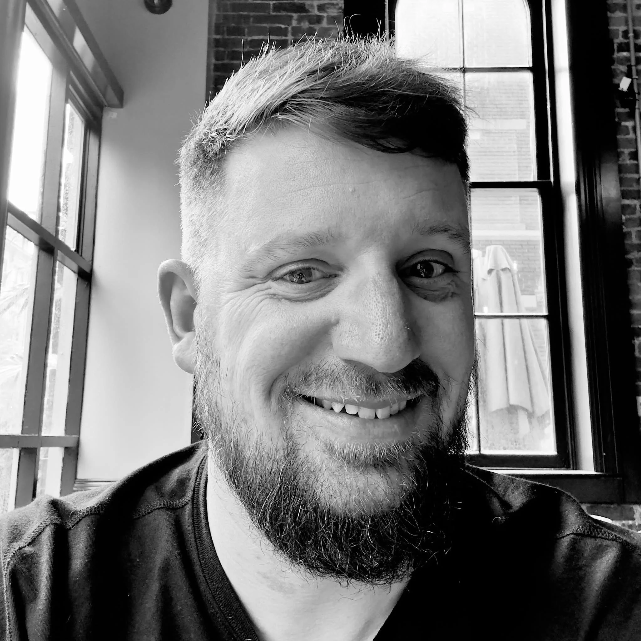 A smiling man with a beard, wearing a dark shirt, sitting near a window in a brick-walled room, with sunlight coming through.