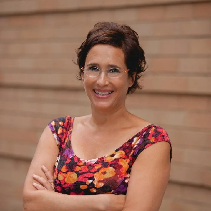 woman with short brown hair, glasses, and a floral top standing in front of a brick wall with arms crossed