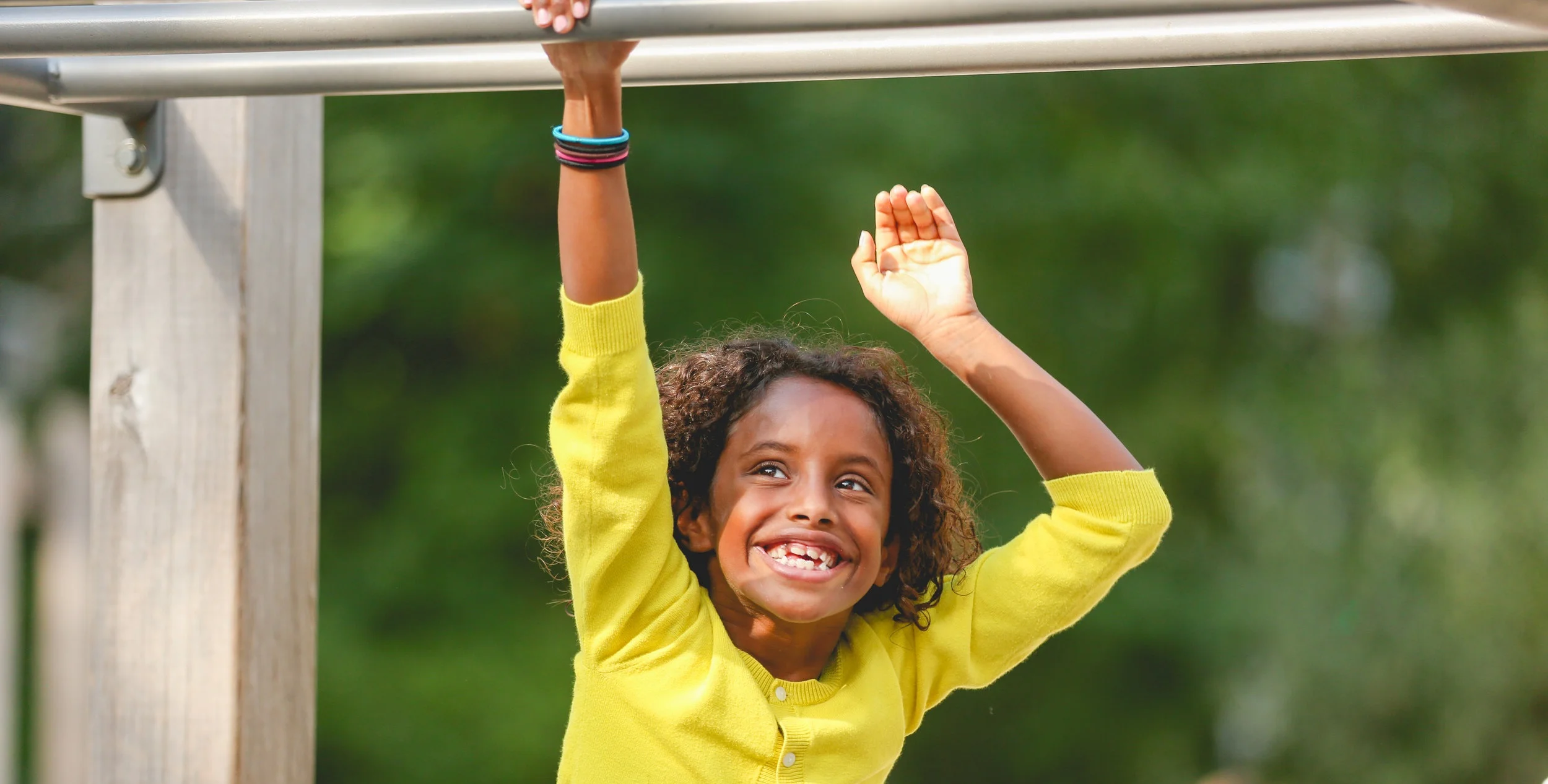 young girl in yellow sweater smiling while playing on monkey bars with green nature background