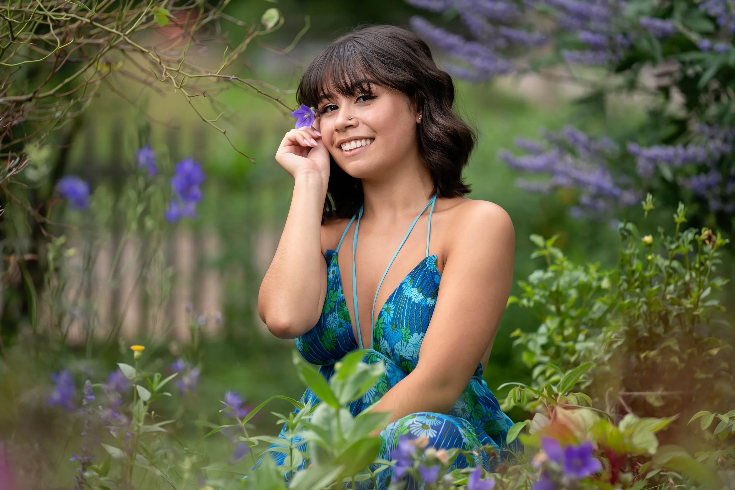 A young woman with dark wavy hair and light to medium skin tone, wearing a blue floral dress, smiling while holding a purple flower to her face in a garden with purple and yellow flowers around her.
