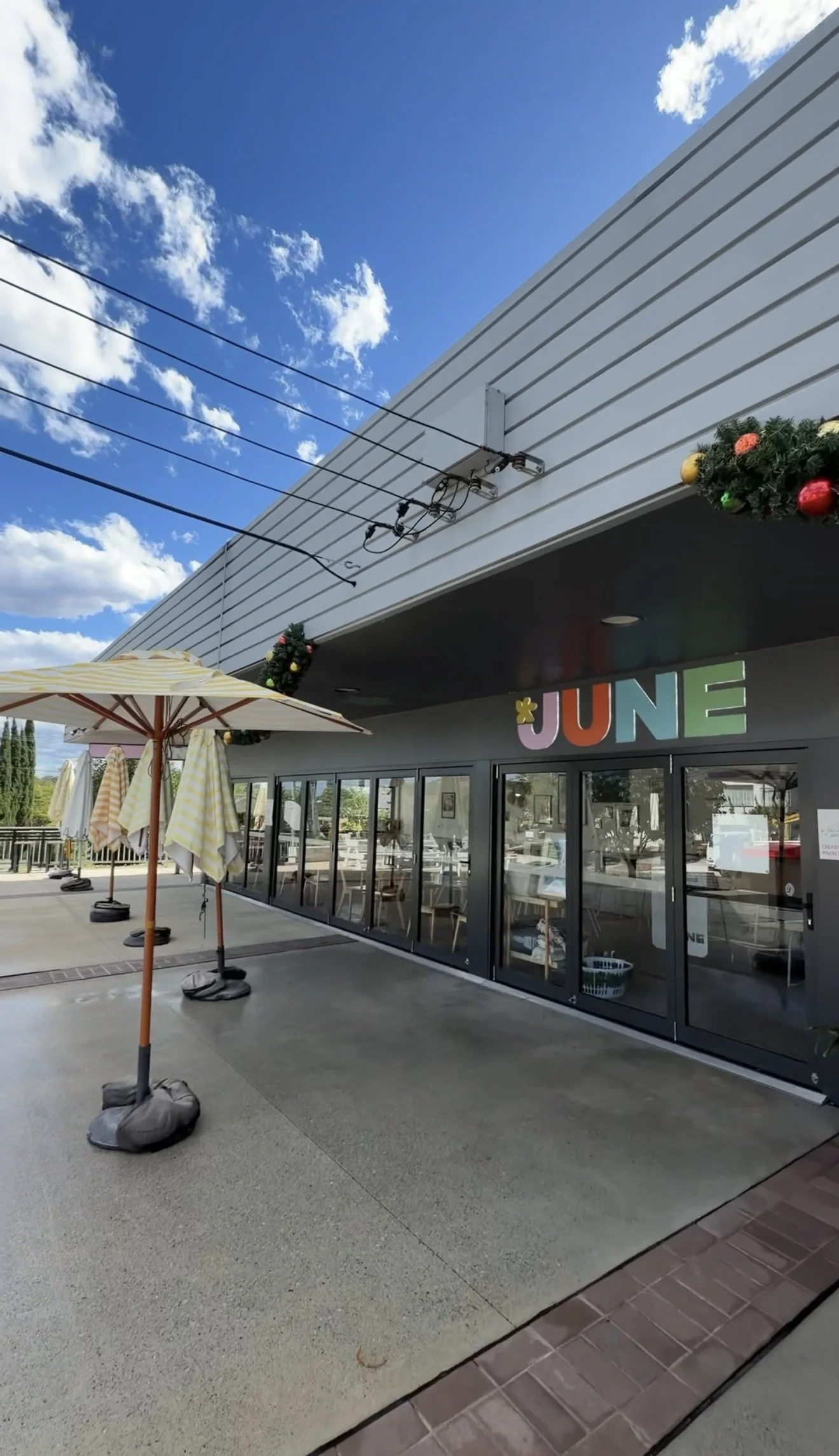 Store storefront decorated for June with colorful letters spelling 'JUNE' above glass doors, with umbrellas and outdoor seating area on a sunny day.