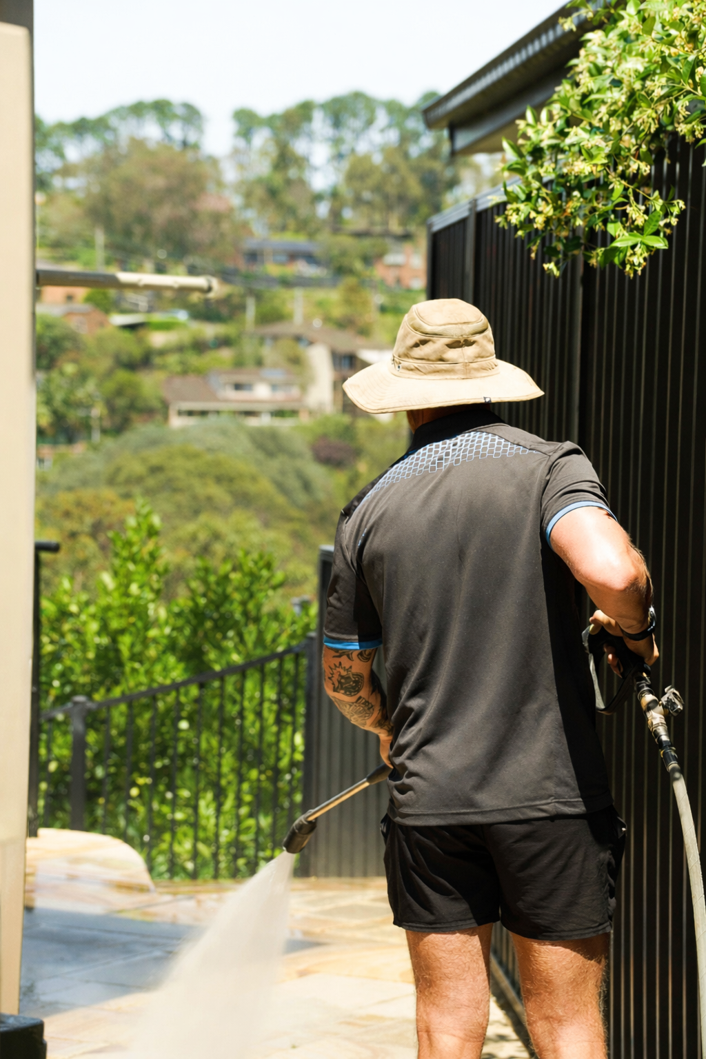 A man wearing a wide-brimmed hat, black shorts, and a black shirt is pressure washing a black metal fence on a sunny day.