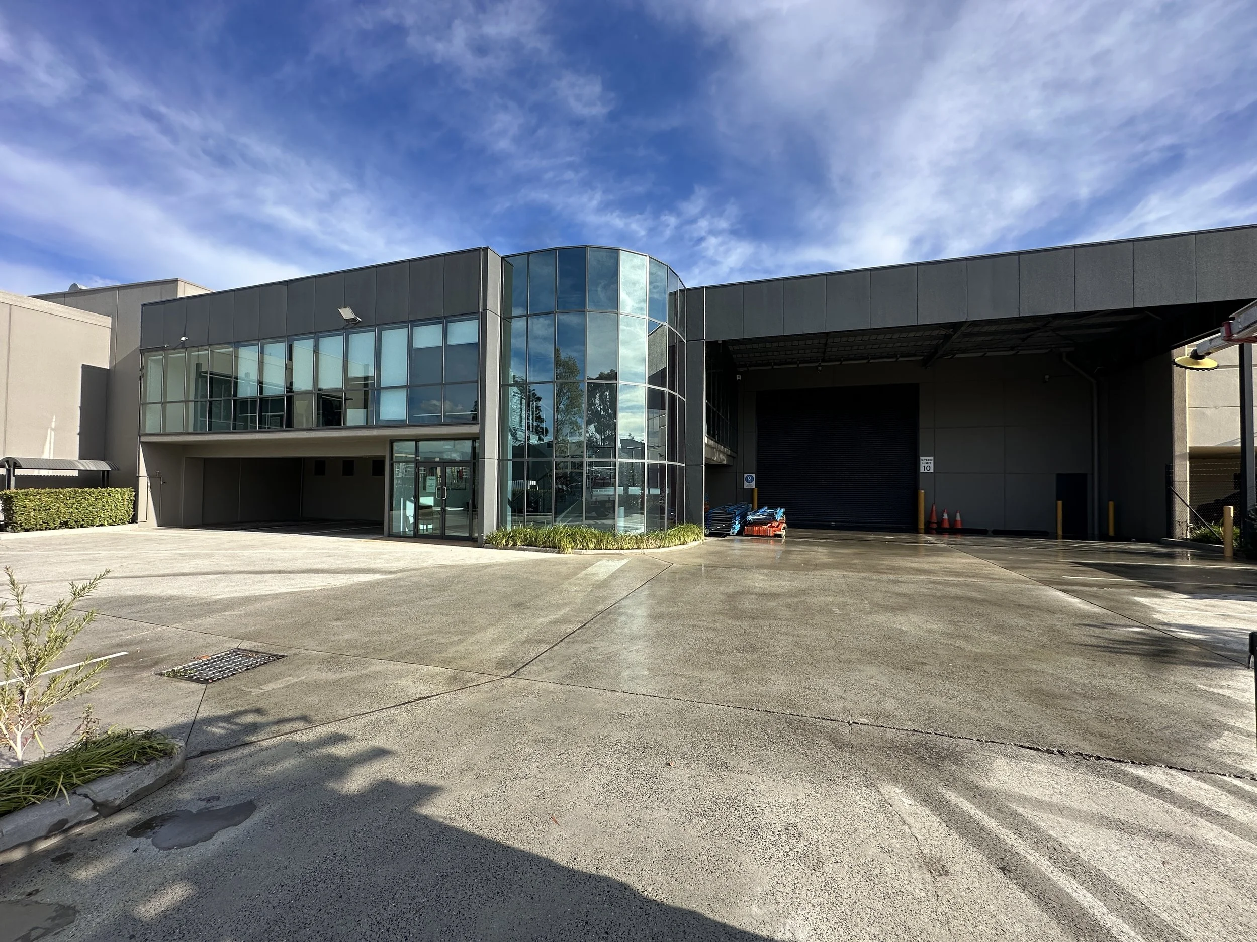 An empty green outdoor tennis court surrounded by trees and a black fence, with cloudy sky overhead.