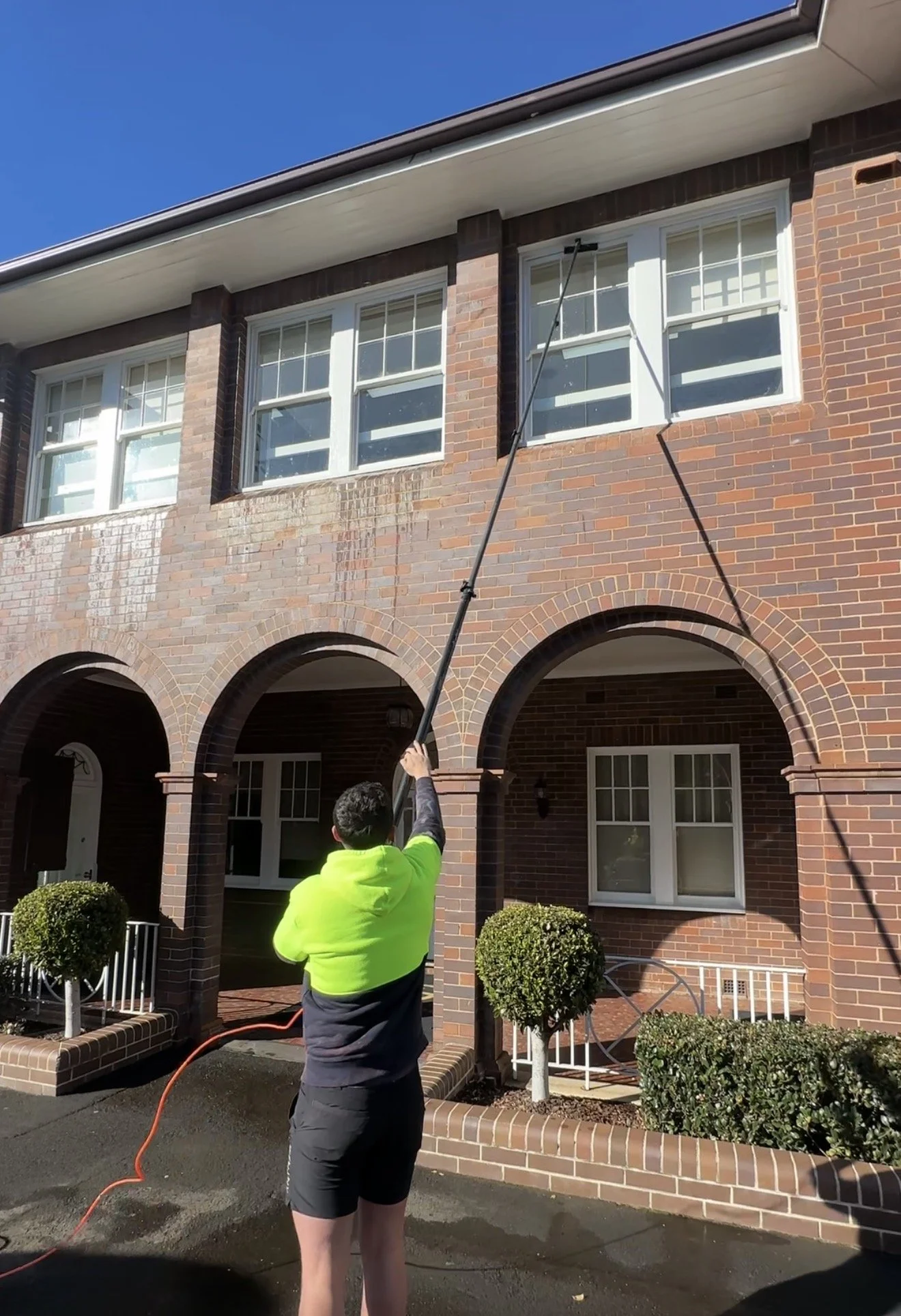 A person in a bright green and black jacket cleans a second-story window using an extendable pole, standing outside a brick townhouse with arched entryways and small landscaped bushes.