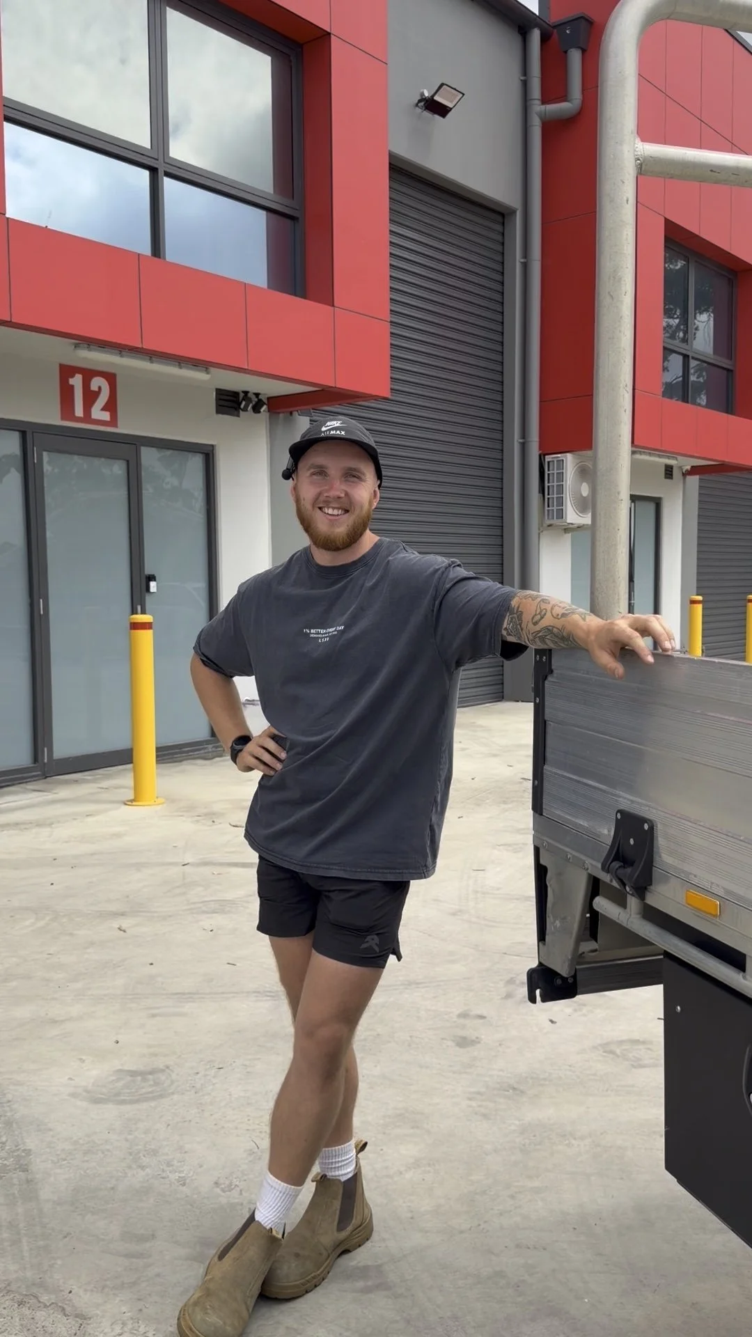 A man smiling and standing outdoors in front of a modern building with red accents, wearing a black cap, gray t-shirt, black shorts, white socks, and tan boots, with his right arm resting on a metal cart.