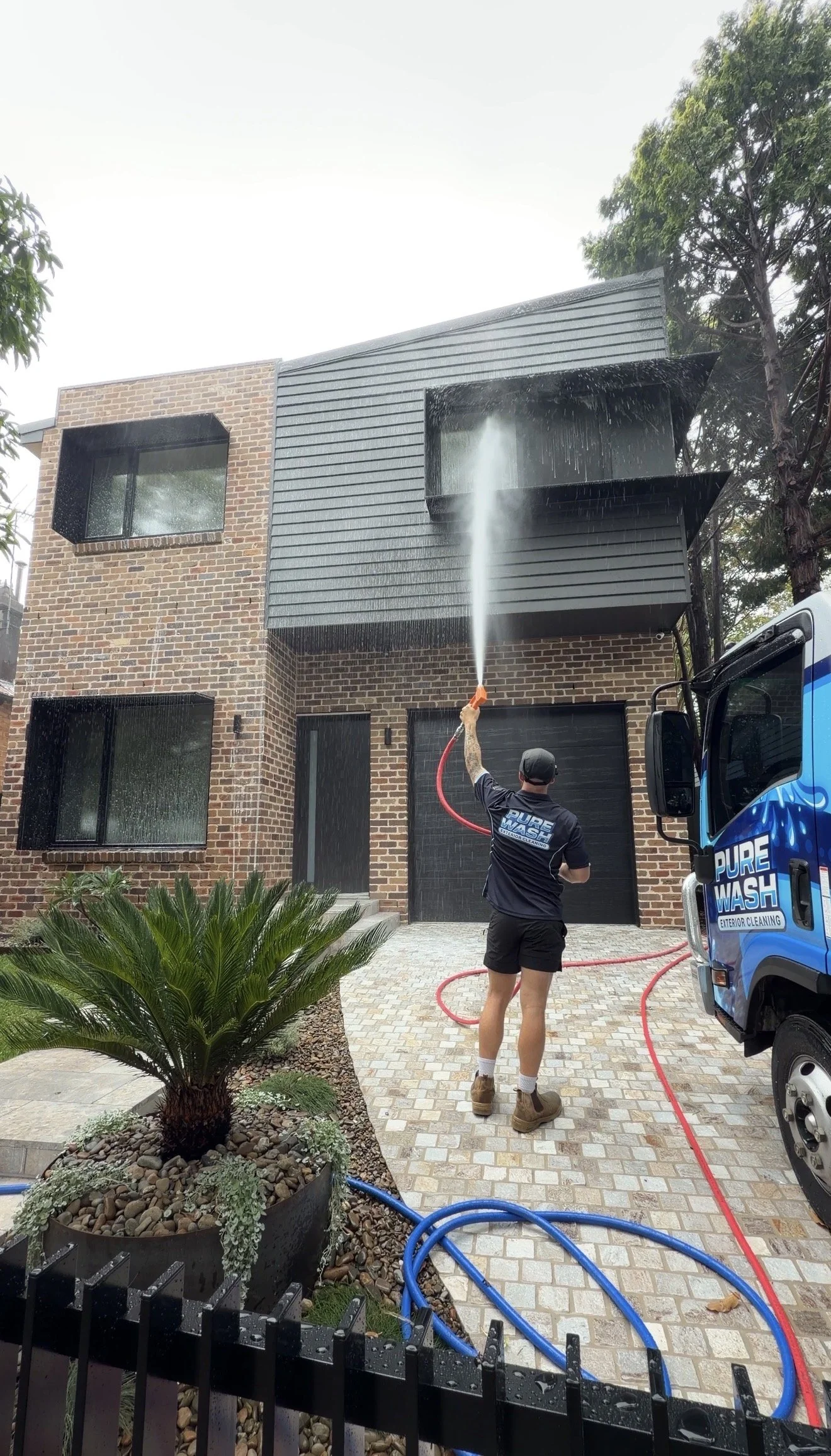 Person wearing a neon yellow safety jacket and shorts using a hose to spray water on a modern two-story house with brick and panel exterior, and a closed black garage door. Overcast sky.