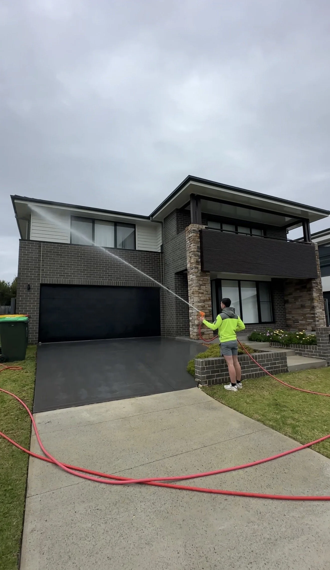 Person wearing a neon yellow safety jacket and shorts using a hose to spray water on a modern two-story house with brick and panel exterior, and a closed black garage door. Overcast sky.