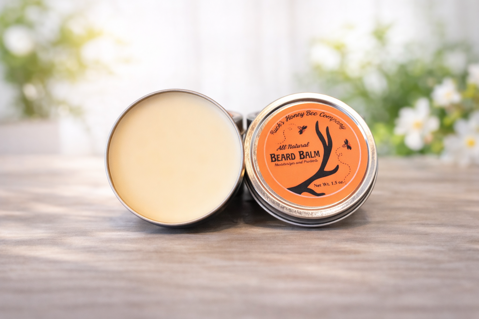 A tin of beard balm with an orange label featuring black graphics of a beard and bees, placed on a wooden surface with blurred green and white background.